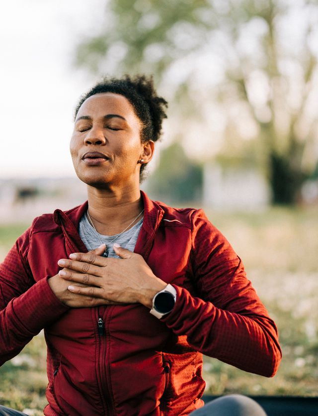 A person wearing a red jacket sits outdoors on grass with eyes closed, hands placed on their chest, appearing calm and relaxed—possibly meditating, practicing mindfulness, or focusing on mental health and wellness.