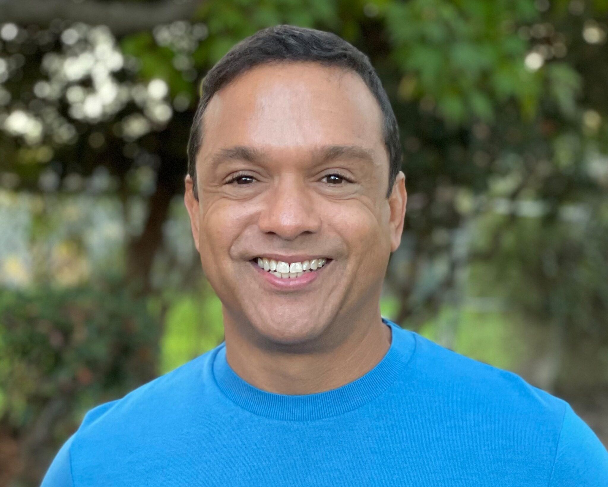 Kenny Williams, with short dark hair and a bright blue sweater, is smiling outdoors amid blurred green trees, embodying someone choosing to be of service.