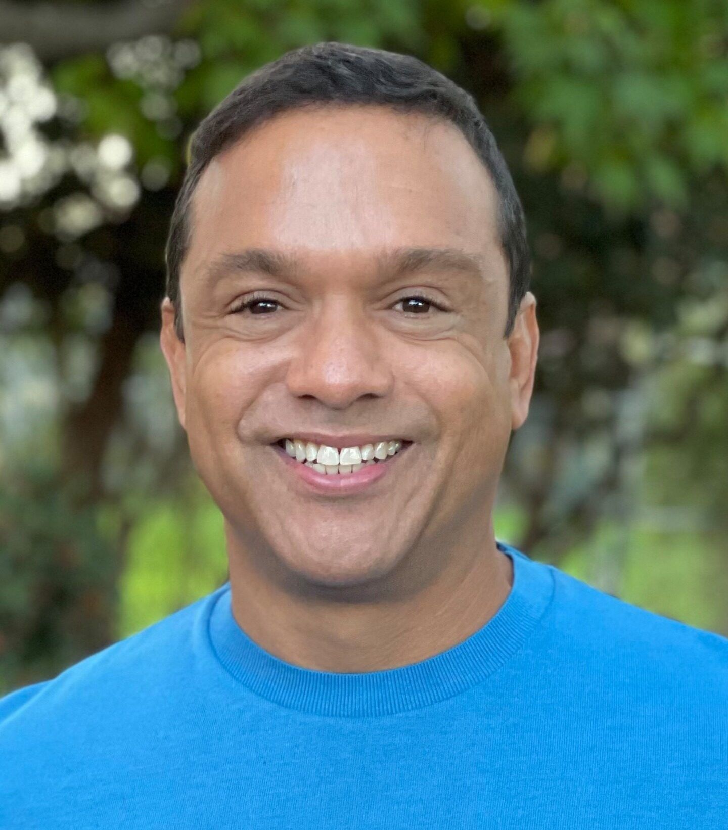 Kenny Williams, with short dark hair and a bright blue sweater, is smiling outdoors amid blurred green trees, embodying someone choosing to be of service.
