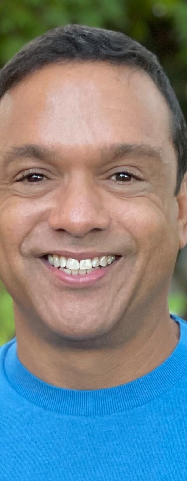 Kenny Williams, with short dark hair and a bright blue sweater, is smiling outdoors amid blurred green trees, embodying someone choosing to be of service.