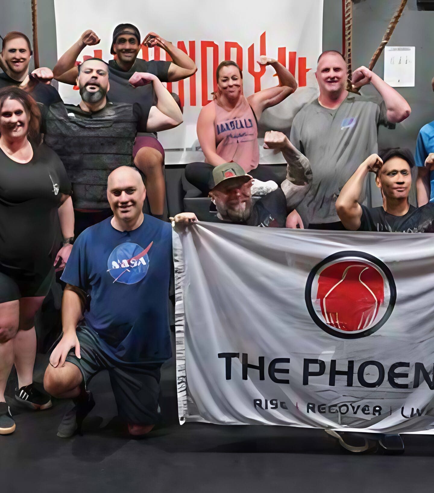 A diverse community at a gym poses together, flexing their arms and smiling. Two people kneel in front holding a banner that reads The Phoenix: Rise Recover Live, celebrating sober active living. Gym equipment is visible in the background.