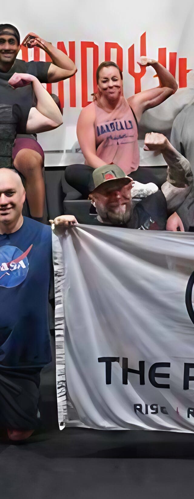 A diverse community at a gym poses together, flexing their arms and smiling. Two people kneel in front holding a banner that reads The Phoenix: Rise Recover Live, celebrating sober active living. Gym equipment is visible in the background.