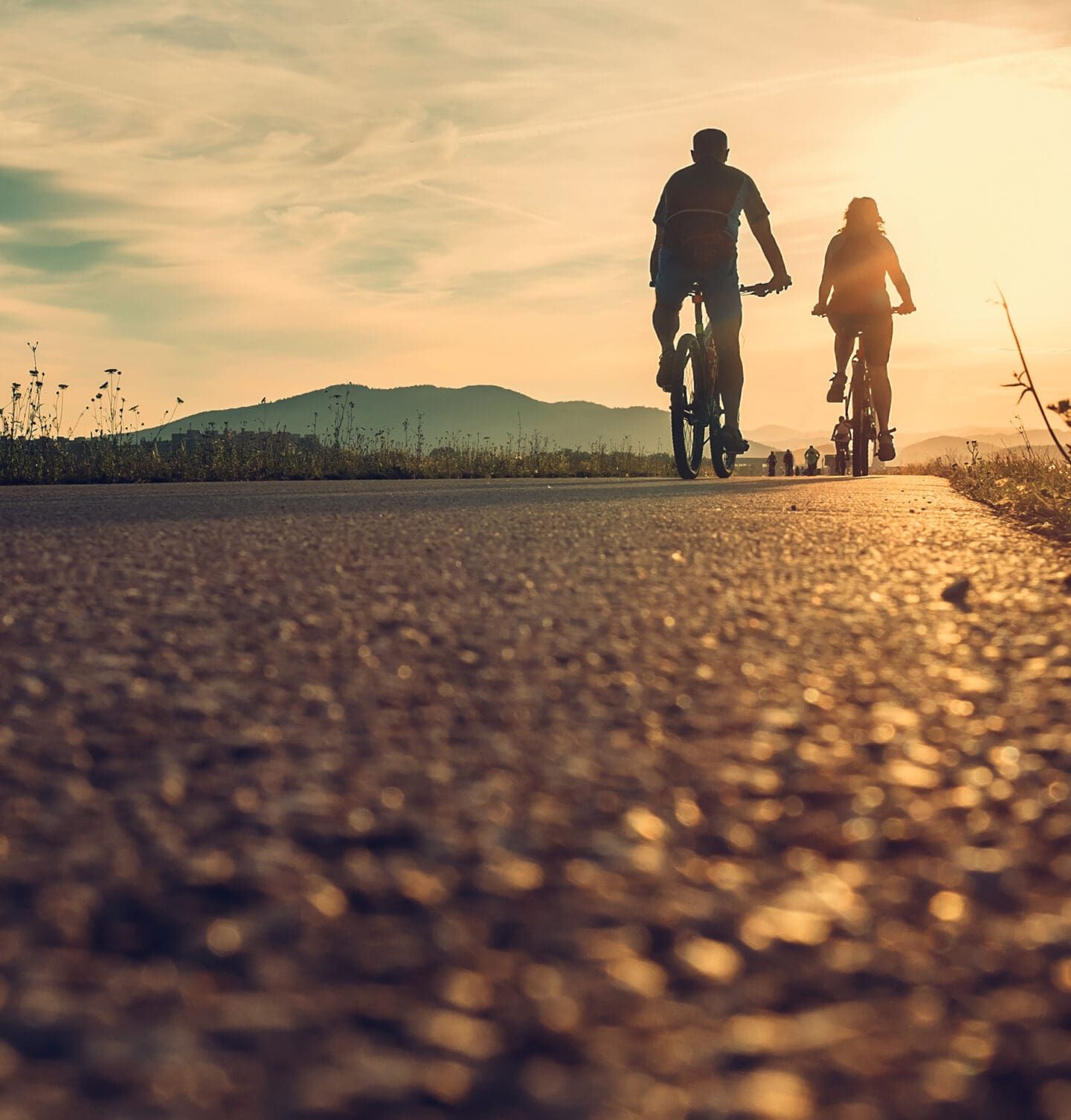 Two people ride bicycles on a paved path at sunset, their silhouettes visible against the bright sky and distant mountains—a peaceful moment that highlights recovery and supports physical well-being.