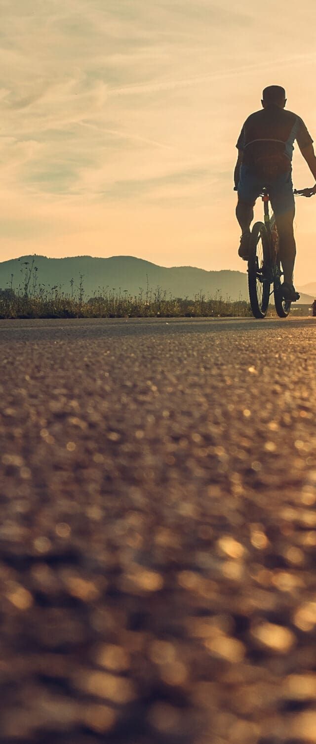 Two people ride bicycles on a paved path at sunset, their silhouettes visible against the bright sky and distant mountains—a peaceful moment that highlights recovery and supports physical well-being.