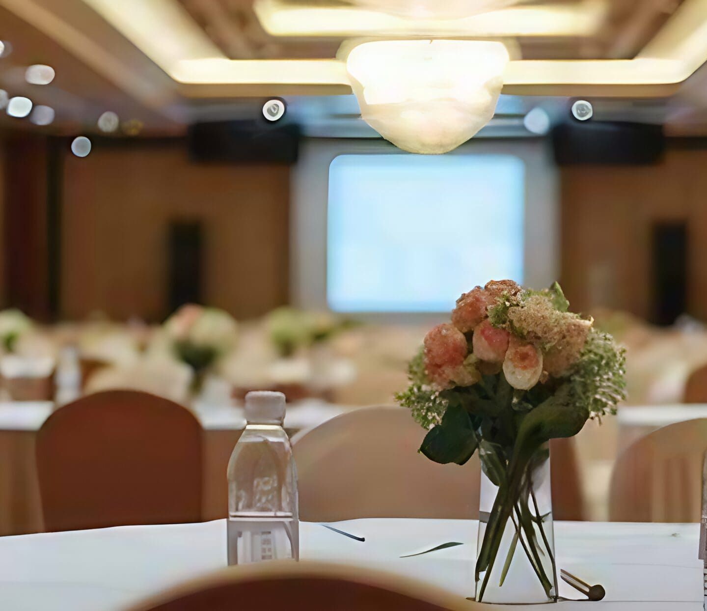 A banquet hall designed for inclusive gatherings, featuring round tables with white cloths, floral centerpieces, and water bottles. Soft lighting fills the room, with a large screen up front and chairs arranged for a welcoming atmosphere.
