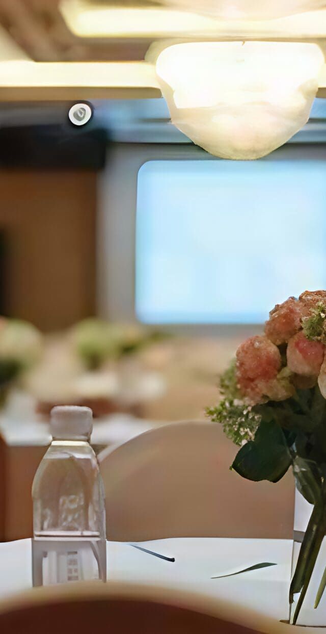 A banquet hall designed for inclusive gatherings, featuring round tables with white cloths, floral centerpieces, and water bottles. Soft lighting fills the room, with a large screen up front and chairs arranged for a welcoming atmosphere.