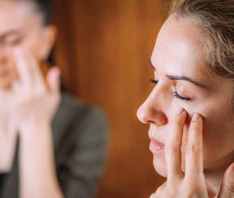Two women with closed eyes gently touch the skin under their eyes with their fingers, appearing to practice EFT Tapping or a relaxation routine in a softly lit indoor setting to help themselves feel better.