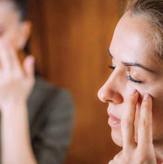 Two women with closed eyes gently touch the skin under their eyes with their fingers, appearing to practice EFT Tapping or a relaxation routine in a softly lit indoor setting to help themselves feel better.