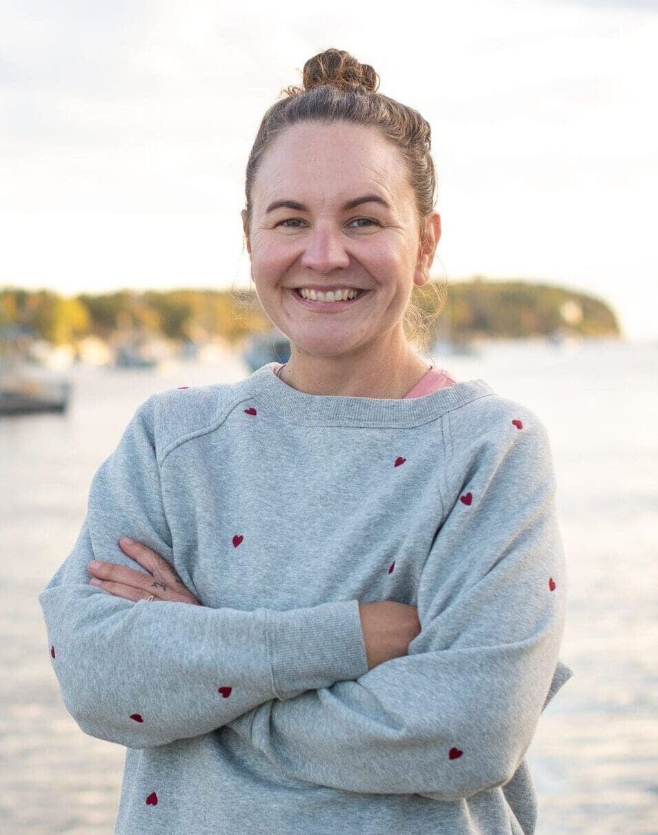 A woman with a top bun and gray sweater stands smiling with arms crossed by a calm waterfront, with boats and a forested shoreline in the background under a partly cloudy sky.