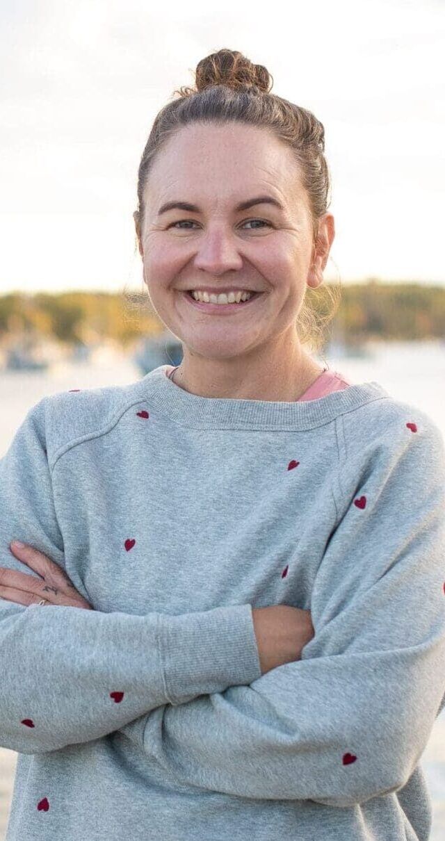 A woman with a top bun and gray sweater stands smiling with arms crossed by a calm waterfront, with boats and a forested shoreline in the background under a partly cloudy sky.
