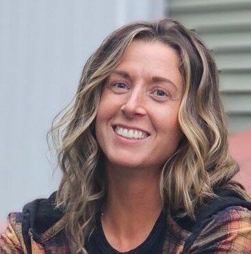 A woman with wavy, light brown hair is smiling at the camera. She is wearing a black shirt and a plaid flannel jacket. The background shows a blurred outdoor setting with a building.