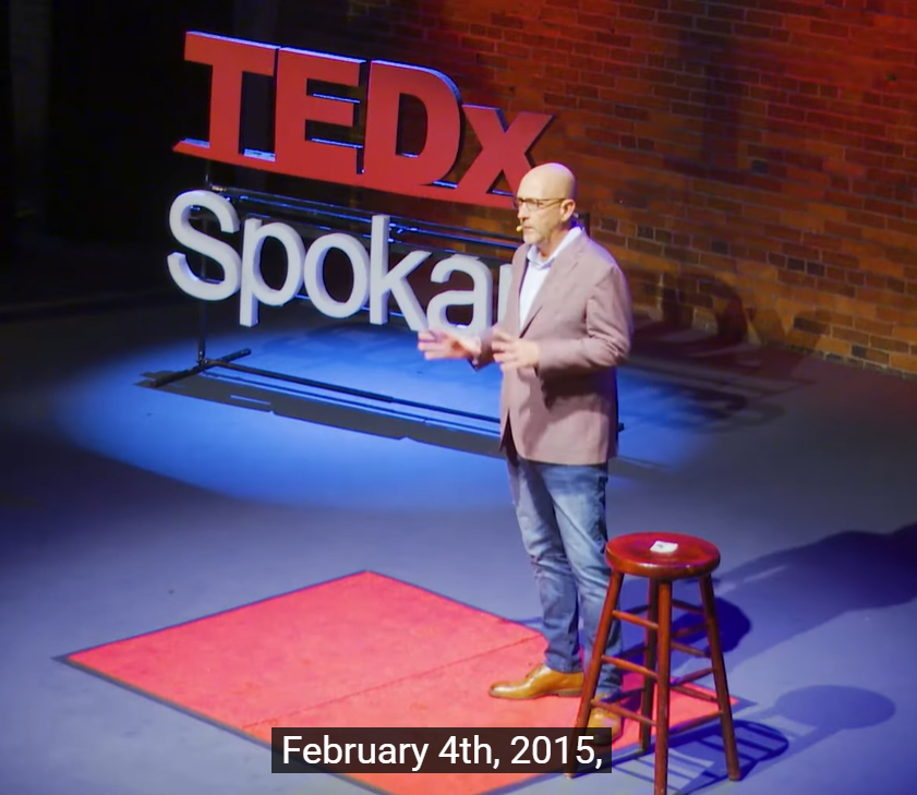 A man stands on stage speaking at a TEDxSpokane event. He is wearing a blazer, jeans, and brown shoes, with a small red carpet and a wooden stool beside him. The subtitle reads, February 4th, 2015,.