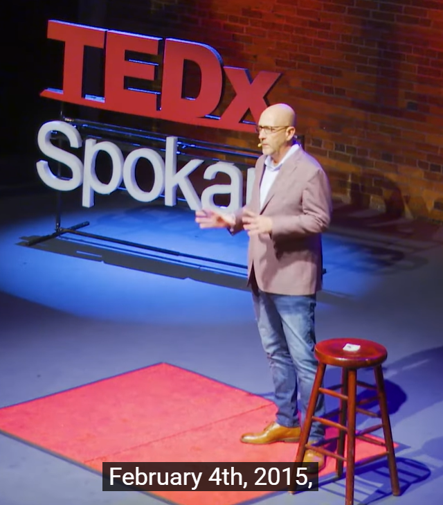 A man stands on stage speaking at a TEDxSpokane event. He is wearing a blazer, jeans, and brown shoes, with a small red carpet and a wooden stool beside him. The subtitle reads, February 4th, 2015,.