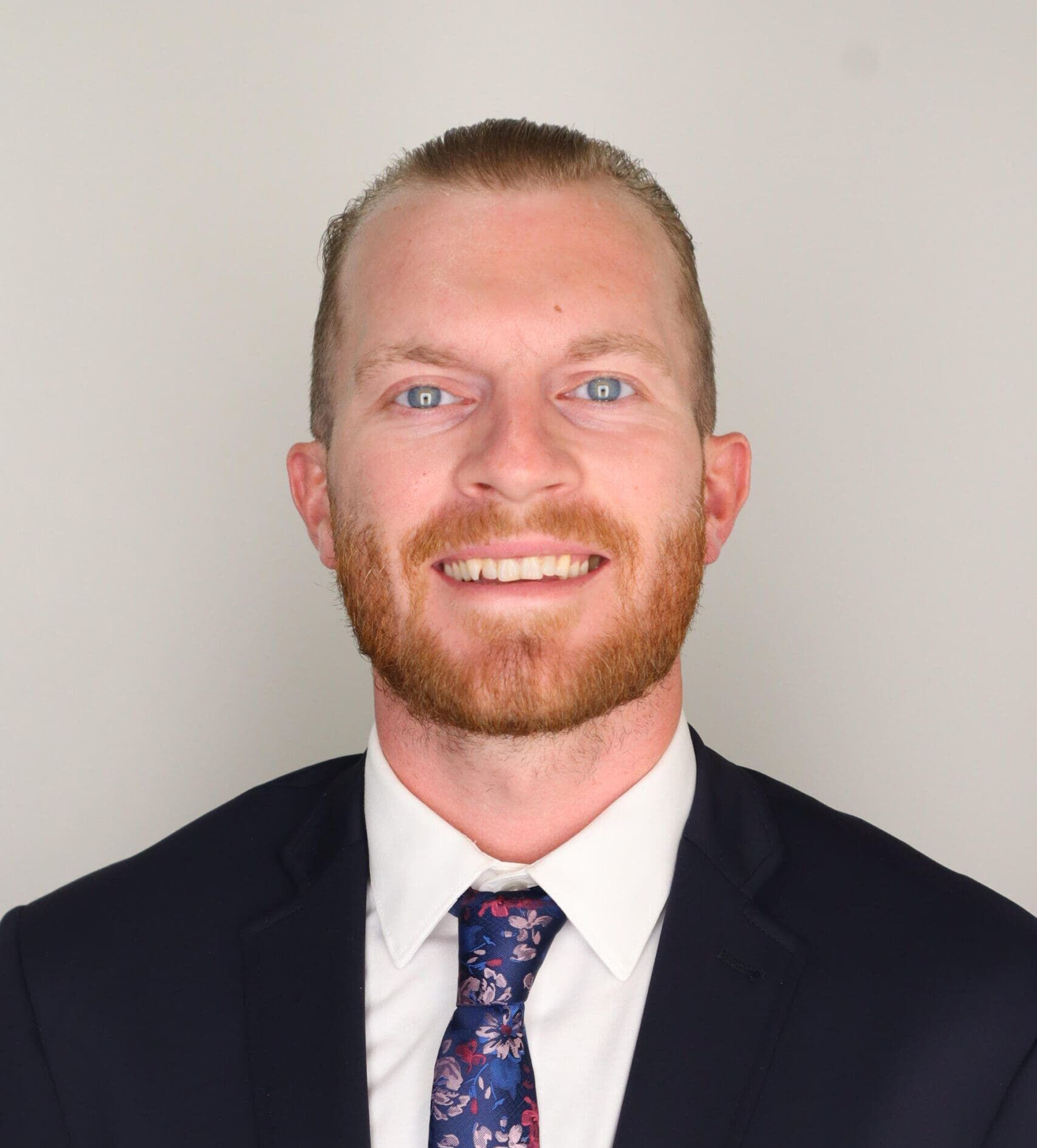 Man with reddish-blonde beard wearing black blazer, white shirt, and blue floral tie smiles at camera against white background.