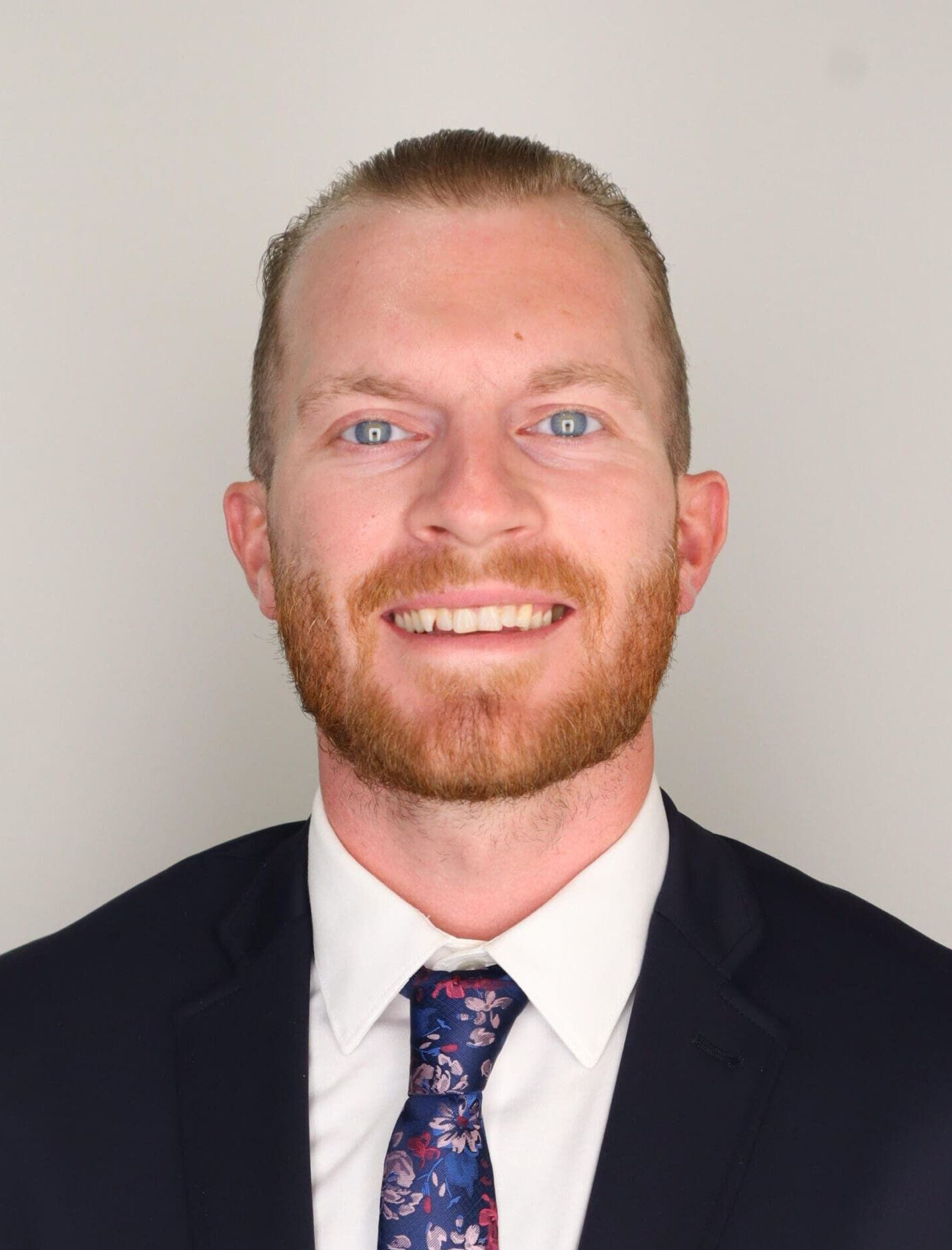 Man with reddish-blonde beard wearing black blazer, white shirt, and blue floral tie smiles at camera against white background.