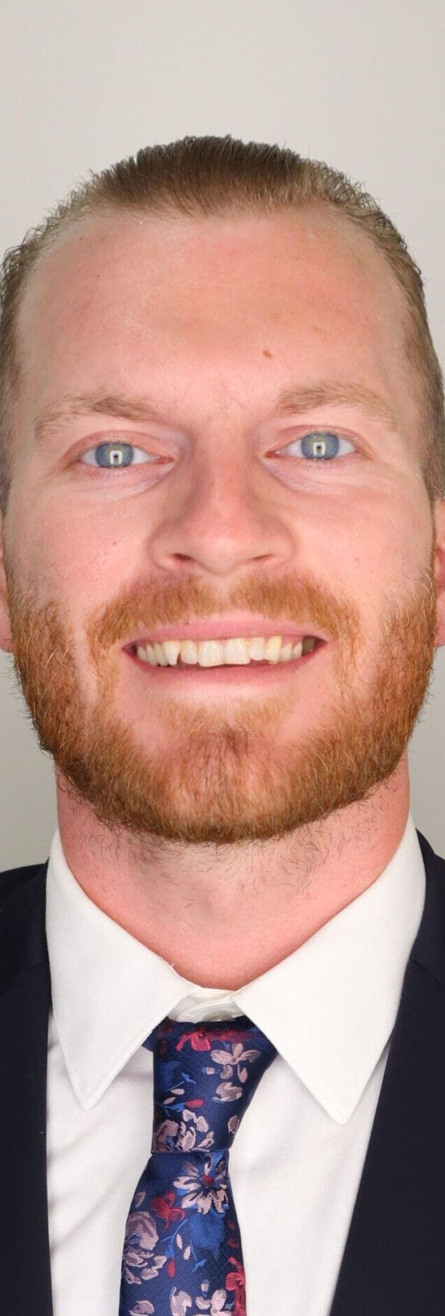 Man with reddish-blonde beard wearing black blazer, white shirt, and blue floral tie smiles at camera against white background.