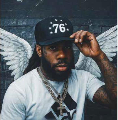 Man wearing "76" baseball cap and white t-shirt poses against blue wall with painted white wings.
