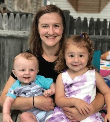 Ashley Reny smiling outdoors with two young children, a baby and a toddler, in front of a wooden fence.