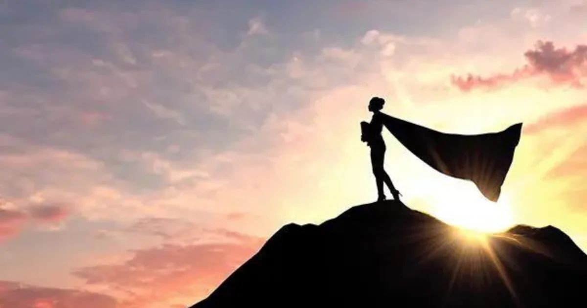 The silhouette of a person in a billowing cape standing on a mountain summit at sunset.