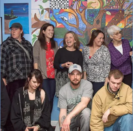 Ten members of the Portland Recovery Community Center standing and seated together in front of a brightly painted mural.