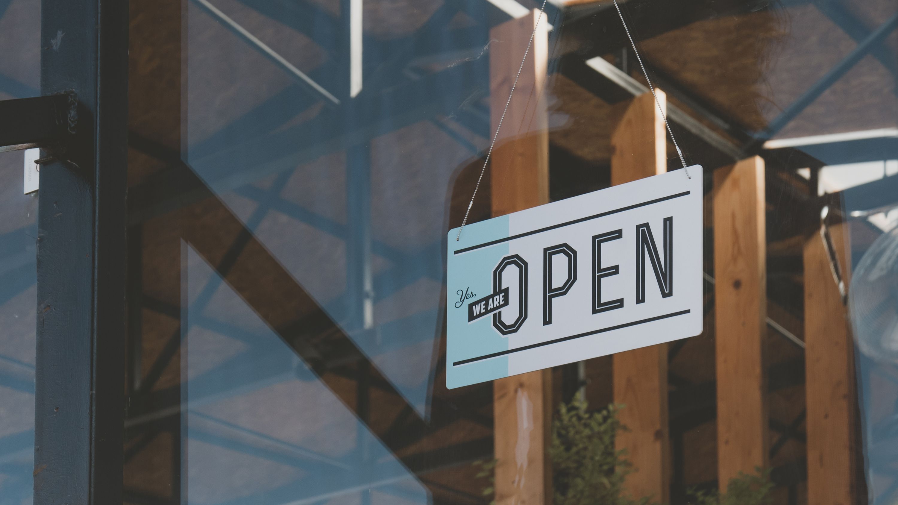 A "Yes, we are OPEN" sign hanging in the window of a business, with wooden posts and steel beams visible through the glass.