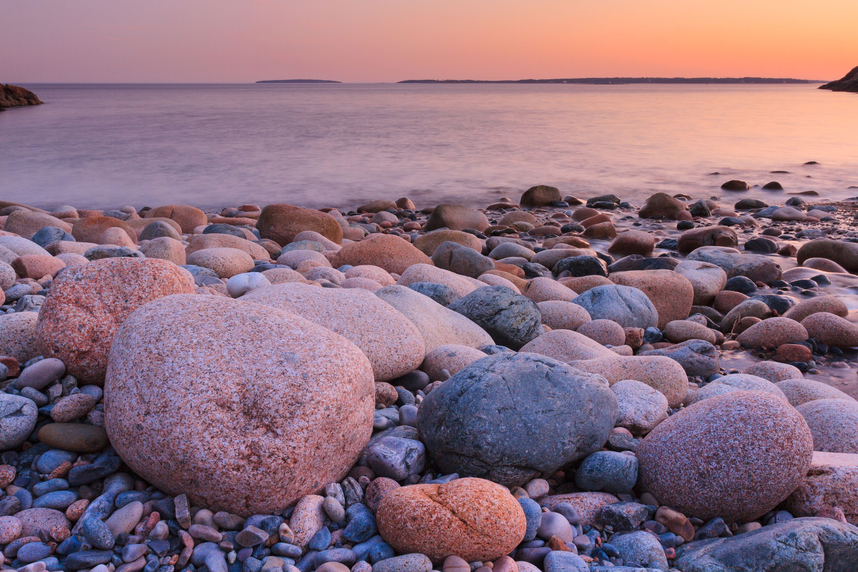Smooth granite stones on a Maine beach at sunset, the tide washing in and the sky glowing pink and orange above a distant headland.