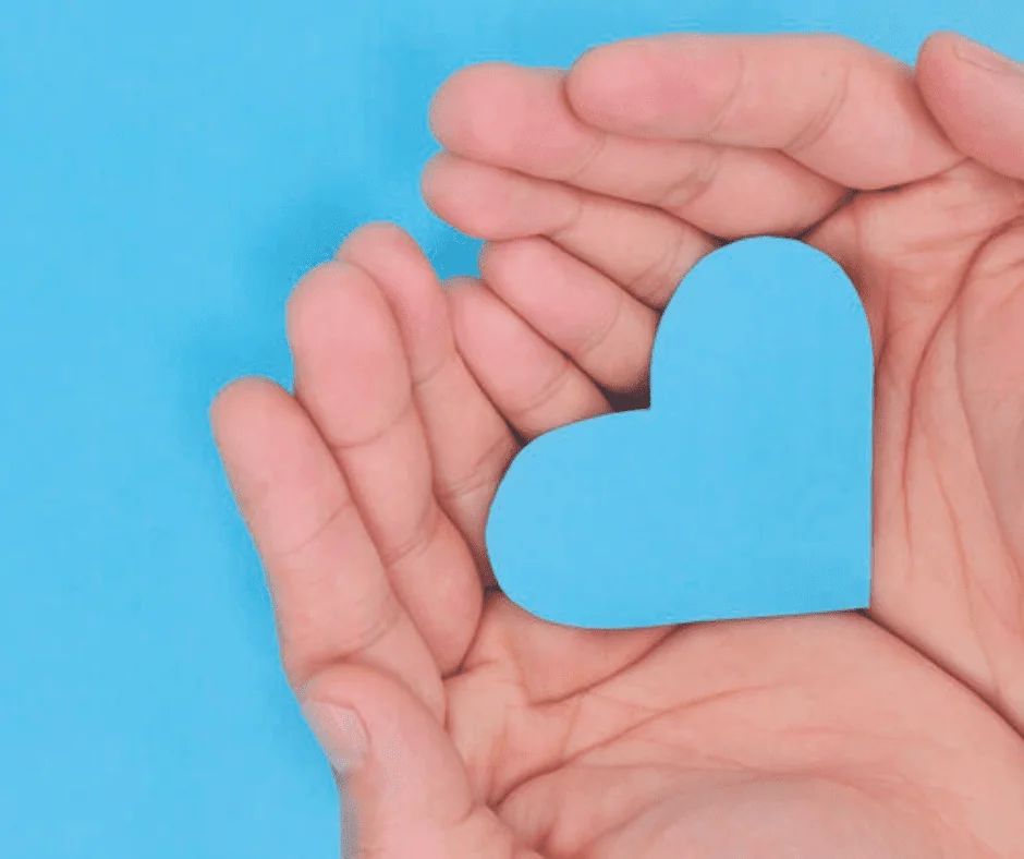 Two cupped hands holding a blue paper heart against a blue background.