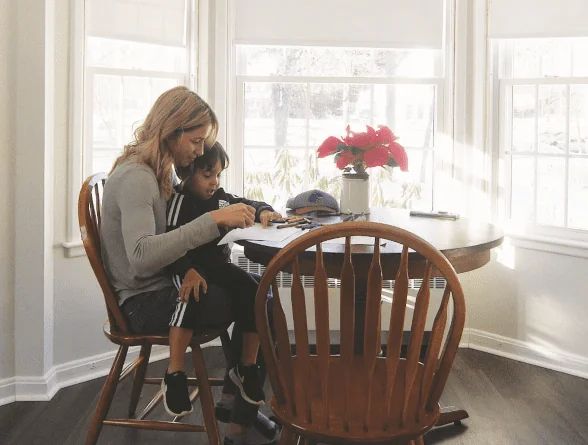 A woman and a child sitting together at a dining table beside bright windows.