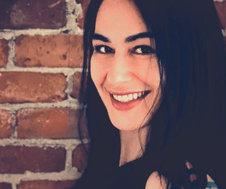 Portrait of Veronica Ingrao, laughing, in front of an exposed brick wall.
