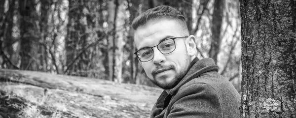Man wearing glasses and a hoodie sits against a tree in a forest setting.