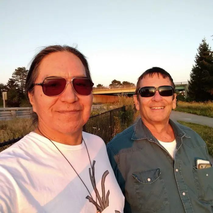 Two people wear sunglasses while standing outdoors near a wooden railing, with trees and fields visible behind them.