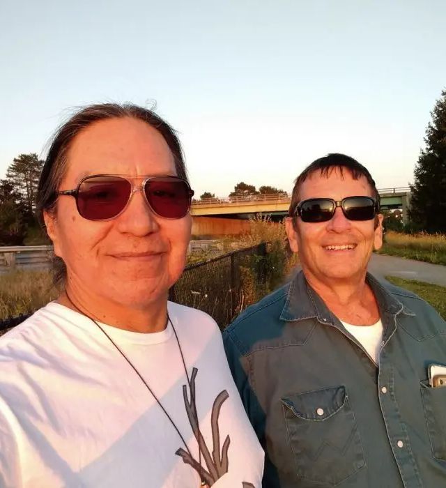 Two people wear sunglasses while standing outdoors near a wooden railing, with trees and fields visible behind them.