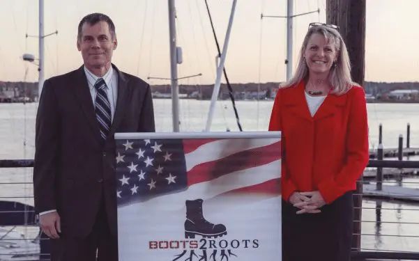 Man in dark suit and woman in red jacket stand beside Boots2Roots banner at waterfront dock with sailboats.
