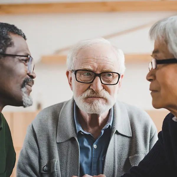 Three people in conversation, one wearing glasses and a gray cardigan, in an indoor setting with wooden walls.