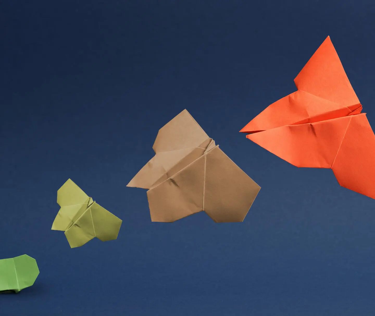 Origami paper shapes in green, yellow, tan, and orange arranged on a dark blue background in increasing sizes.