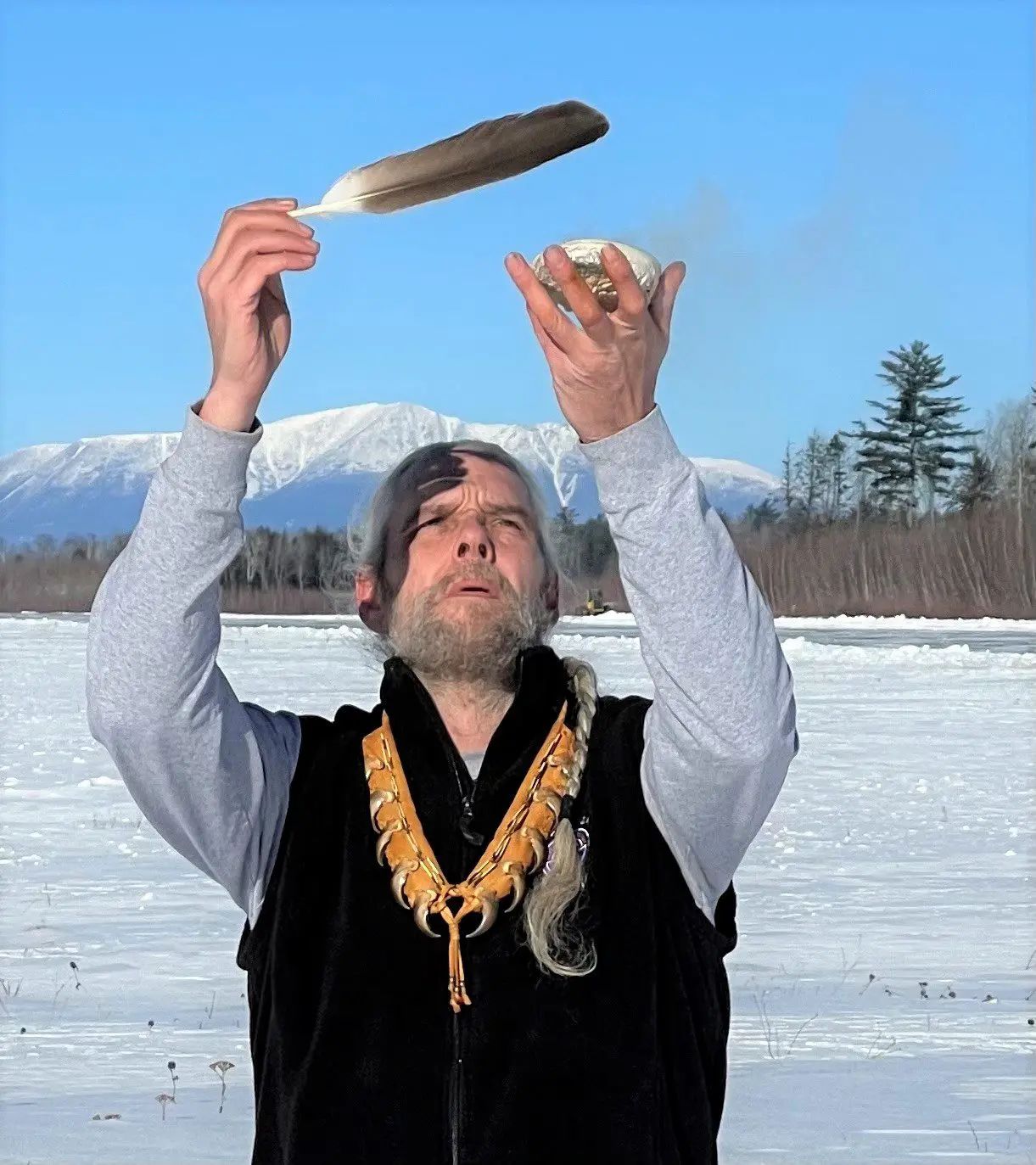 Man holds feather and wrapped item overhead while standing on frozen lake with mountains and evergreen trees behind him.