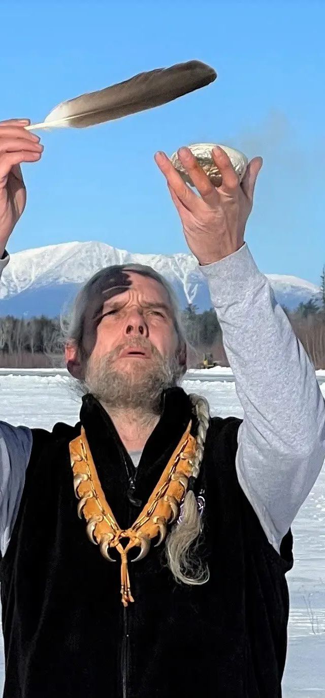 Man holds feather and wrapped item overhead while standing on frozen lake with mountains and evergreen trees behind him.