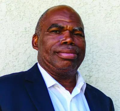 Anthony Brown, a man wearing a dark blazer and light blue shirt, smiles at the camera against a white textured wall.